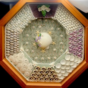 May include: A framed octagonal artwork made of various seashells arranged in a decorative pattern. The centre features a large white seashell with a small cluster of purple and white flowers. The frame is made of light brown wood.