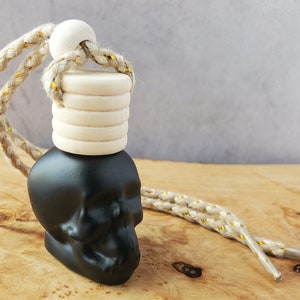 Puede incluir: Ambientador de coche en forma de calavera negra con tapa de madera blanca y cordón beige y dorado.