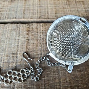 May include: A silver tea infuser with a chain and a bee and honeycomb charm. The infuser is round and has a fine mesh.
