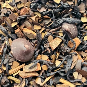 May include: Close-up of loose leaf tea blend with dark brown tea leaves, light brown bark pieces, and two round, dark chocolate-covered pieces. The tea blend has a variety of textures and colors.