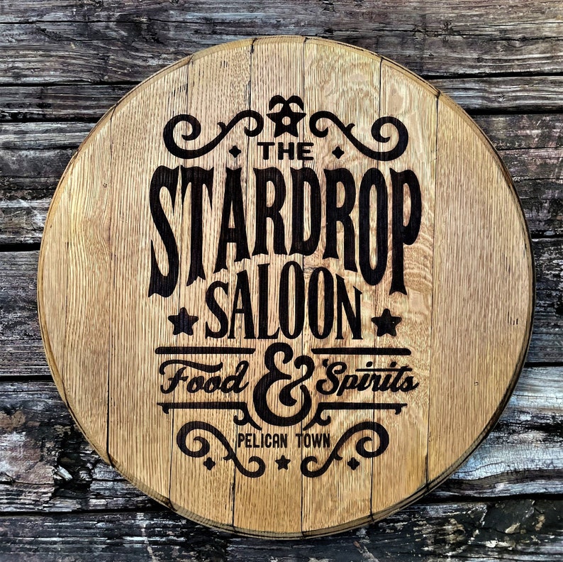 May include: Circular wooden sign with the text "THE STARDROP SALOON" in a bold, vintage font. The sign also includes "Food & Spirits" and "Pelican Town". The sign is made of light brown wood.
