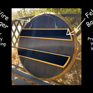 May include: A round, dark wood wall shelf with two black shelves and gold trim. The shelf has a wire hanger for easy hanging and felt backing to protect the wall. The wood has a natural grain, and the overall design is rustic.