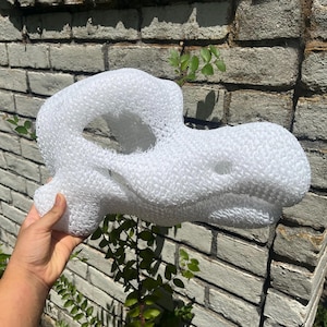 May include: A white, three-dimensional dinosaur skull sculpture crafted from a textured, mesh-like material. The structure features a hollow eye socket, a prominent snout, and a jagged jawline, showcasing a lightweight, porous, and synthetic construction.