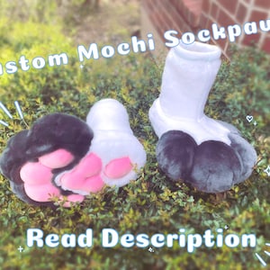 May include: A pair of custom mochi sock paws with black fur and pink paw pads. The sock paws are white and have a fluffy texture. The text "Custom Mochi Sockpaws" is written in a pink font on a white background. The text "Read Description" is written in a blue font on a white background.