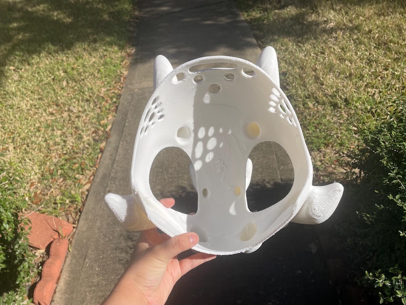 Puede incluir: Una base de m&aacute;scara blanca impresa en 3D con forma redondeada y dos protuberancias prominentes en forma de cuerno en los lados. La estructura incluye grandes aberturas circulares para los ojos y una serie de orificios de ventilaci&oacute;n.