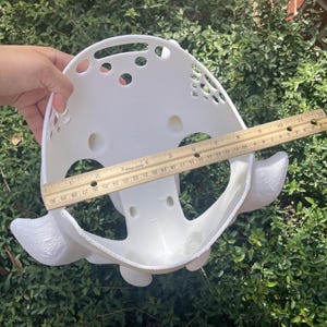 Puede incluir: Una m&aacute;scara blanca impresa en 3D con forma redondeada, m&uacute;ltiples recortes circulares y protuberancias laterales decorativas. Una regla de madera se coloca en el centro, mostrando una anchura de unos 28 cent&iacute;metros de borde a borde.