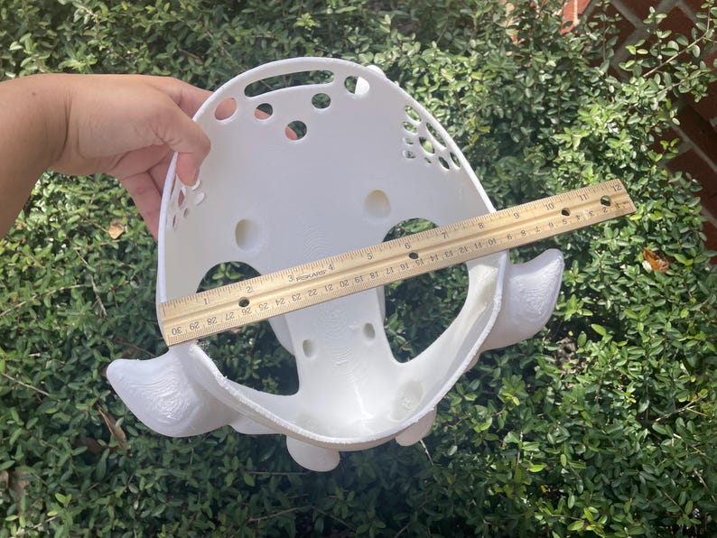 Puede incluir: Una m&aacute;scara blanca impresa en 3D con forma redondeada, m&uacute;ltiples recortes circulares y protuberancias laterales decorativas. Una regla de madera se coloca en el centro, mostrando una anchura de unos 28 cent&iacute;metros de borde a borde.