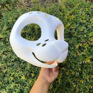 Könnte beinhalten: Eine weiße, 3D-gedruckte Tiermaske mit großen Augenöffnungen, kleiner Nase und einem lächelnden Mund. Die Maske hat mehrere kleine Löcher oben und wird vor einem Hintergrund aus grünem Laub gehalten.