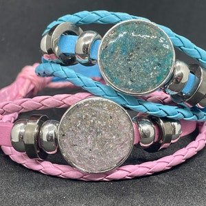 Könnte beinhalten: Zwei geflochtene Lederarmbänder mit silbernen Metallperlen und runden Harz-Anhängern. Ein Armband ist hellblau und das andere ist rosa.