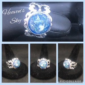 Könnte beinhalten: Ein silberner Ring mit einem blauen und weißen glitzernden Stein, der in einem filigranen Design gefasst ist. Der Ring befindet sich auf einem schwarzen Präsentationsständer. Der Text "Heaven's Sky" ist im Bild sichtbar.