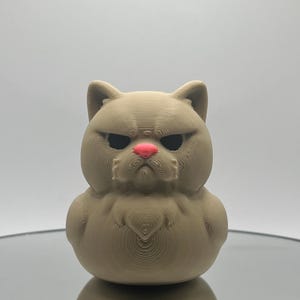 May include: A 3D printed figurine of a brown cat with a grumpy expression. The cat has a pink nose and black eyes.