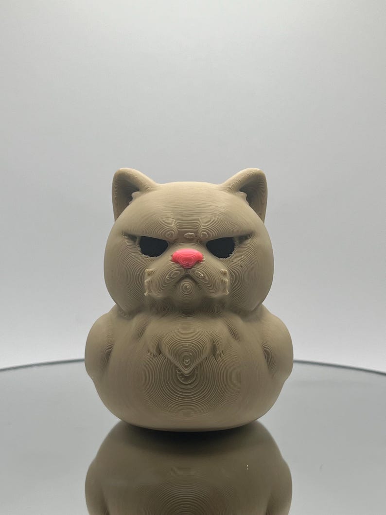 May include: A 3D printed figurine of a brown cat with a grumpy expression. The cat has a pink nose and black eyes.