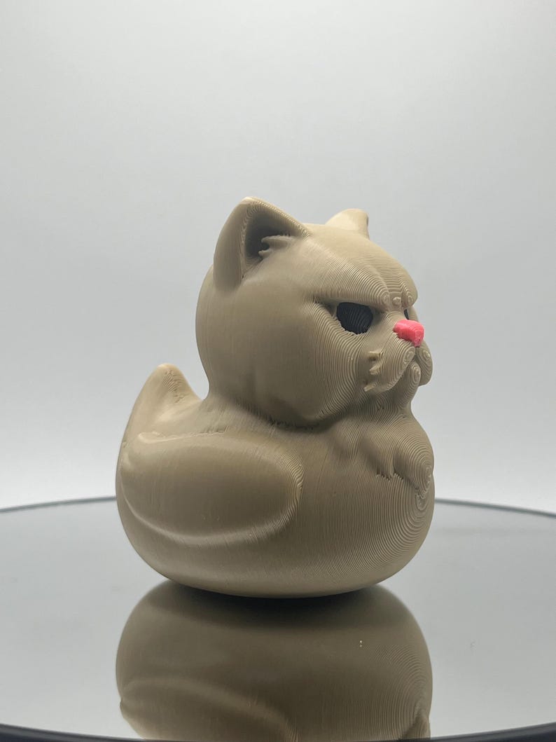 May include: A tan rubber duck with a grumpy cat face. The duck has a pink nose and black eyes.