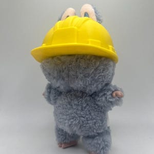 May include: A soft, grey toy animal wearing a yellow hard hat. The toy has small pink hands and feet, and blue ears are visible. The toy is set against a plain white backdrop.