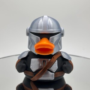 May include: A silver and black rubber ducky with an orange beak and a Mandalorian helmet.