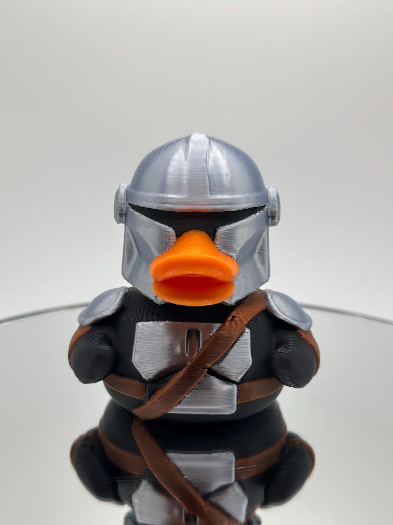 May include: A silver and black rubber ducky with an orange beak and a Mandalorian helmet.