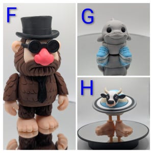 Puede incluir: Tres figuras caprichosas: un personaje marr&oacute;n parecido a Pie Grande con sombrero de copa y gafas de sol, un delf&iacute;n gris con zapatillas azules y una vaca en un plato con patas. Cada figura es detallada y &uacute;nica.