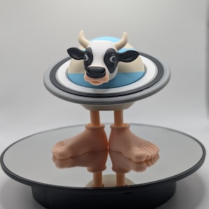 Puede incluir: Una caprichosa figura de vaca con cara blanca y negra, cuernos y grandes orejas negras. La vaca est&aacute; posada sobre una plataforma circular con un dise&ntilde;o de rayas azules y blancas, sostenida por dos pies. La figura se exhibe sobre una superficie espejada.
