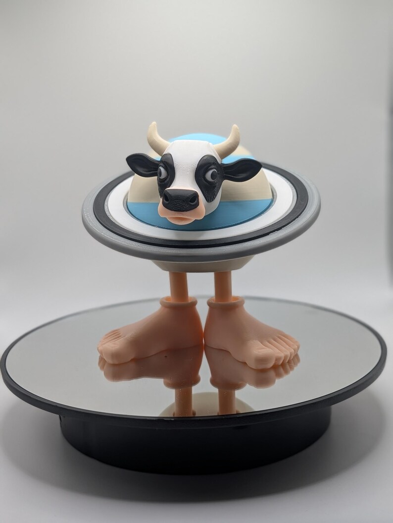 Puede incluir: Una caprichosa figura de vaca con cara blanca y negra, cuernos y grandes orejas negras. La vaca est&aacute; posada sobre una plataforma circular con un dise&ntilde;o de rayas azules y blancas, sostenida por dos pies. La figura se exhibe sobre una superficie espejada.