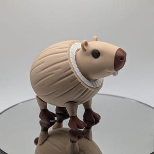 Puede incluir: Una figura de carpincho marr&oacute;n con un collar blanco. La figura tiene patas marrones y una nariz marr&oacute;n. El carpincho est&aacute; de pie sobre una superficie reflectante. La figura es una representaci&oacute;n estilizada del animal.
