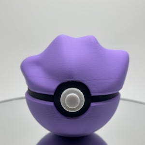 May include: A purple and black Poke Ball with a white button in the centre. The Poke Ball is a popular collectible toy.