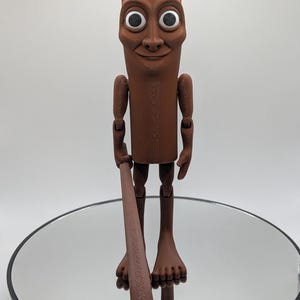 Puede incluir: Una figura articulada marr&oacute;n con una cara sonriente, ojos blancos grandes y un garrote. La figura tiene brazos y piernas articulados y est&aacute; de pie sobre una superficie reflectante. La figura es un dise&ntilde;o &uacute;nico y caprichoso.