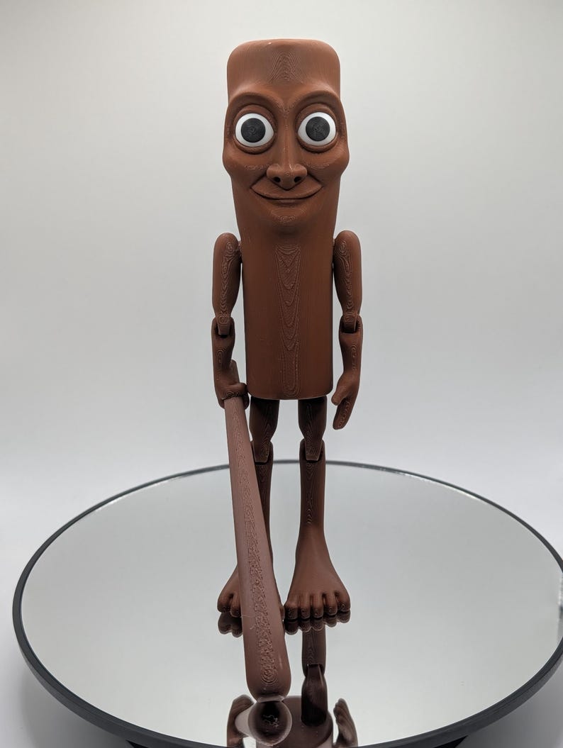 Puede incluir: Una figura articulada marr&oacute;n con una cara sonriente, ojos blancos grandes y un garrote. La figura tiene brazos y piernas articulados y est&aacute; de pie sobre una superficie reflectante. La figura es un dise&ntilde;o &uacute;nico y caprichoso.