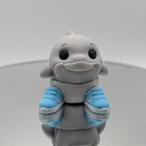 Puede incluir: Una figura gris de un delf&iacute;n con zapatillas de color azul claro. El delf&iacute;n tiene ojos negros y una expresi&oacute;n sonriente. La figura se refleja en una superficie espejada.