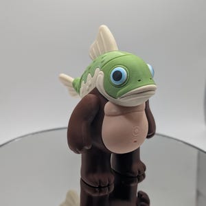 Puede incluir: Una figura caprichosa de una criatura parecida a un pez con cabeza verde, ojos azules y cuerpo marr&oacute;n. La figura tiene vientre y aletas de color beige claro. La figura est&aacute; sobre una superficie reflectante.