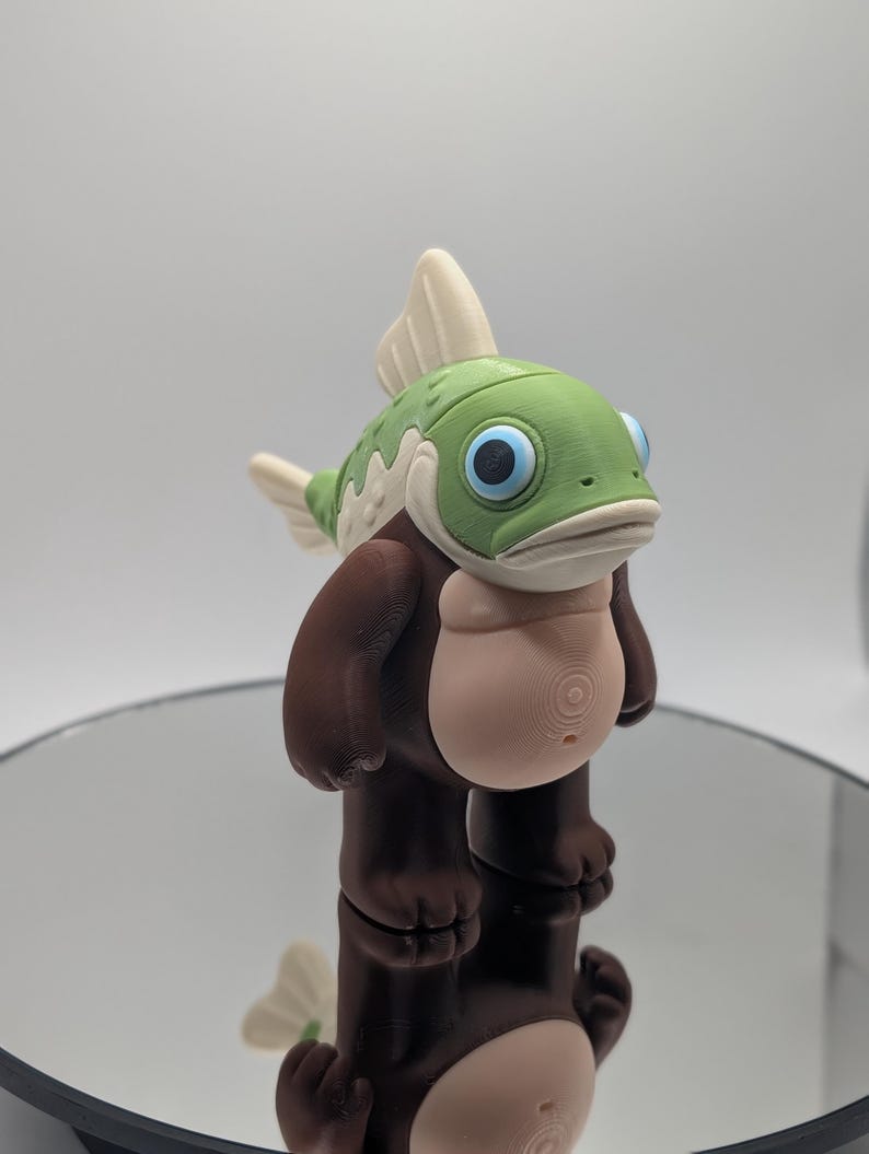 Puede incluir: Una figura caprichosa de una criatura parecida a un pez con cabeza verde, ojos azules y cuerpo marr&oacute;n. La figura tiene vientre y aletas de color beige claro. La figura est&aacute; sobre una superficie reflectante.