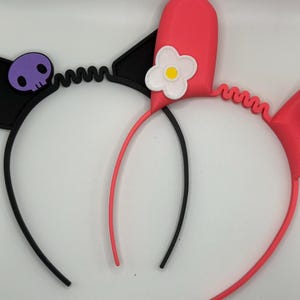 May include: Two black and pink headbands with ears. One headband has a purple skull and the other has a white flower.