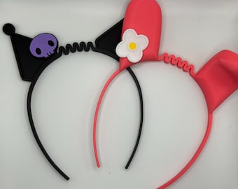 May include: Two black and pink headbands with ears. One headband has a purple skull and the other has a white flower.