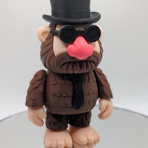 Puede incluir: Una figura caprichosa que se asemeja a un Bigfoot, con un sombrero de copa negro, gafas redondas y una corbata negra. La figura tiene una textura marr&oacute;n similar a la piel, una nariz rosa y est&aacute; de pie sobre una superficie reflectante. El dise&ntilde;o es de dibujos animados.