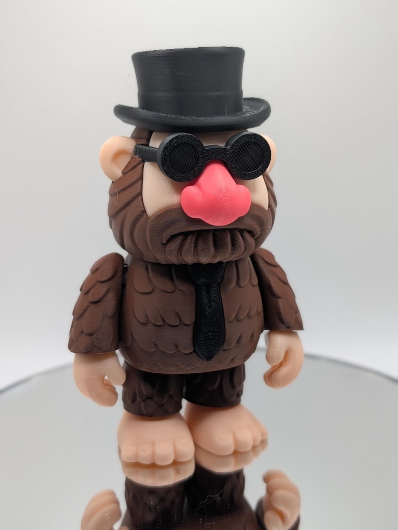 Puede incluir: Una figura caprichosa que se asemeja a un Bigfoot, con un sombrero de copa negro, gafas redondas y una corbata negra. La figura tiene una textura marr&oacute;n similar a la piel, una nariz rosa y est&aacute; de pie sobre una superficie reflectante. El dise&ntilde;o es de dibujos animados.