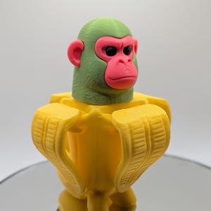 Puede incluir: Una escultura impresa en 3D de un mono con cabeza verde, cara y orejas rosas y ojos negros. El mono lleva un traje amarillo con forma de pl&aacute;tano. La escultura est&aacute; sobre una superficie reflectante.