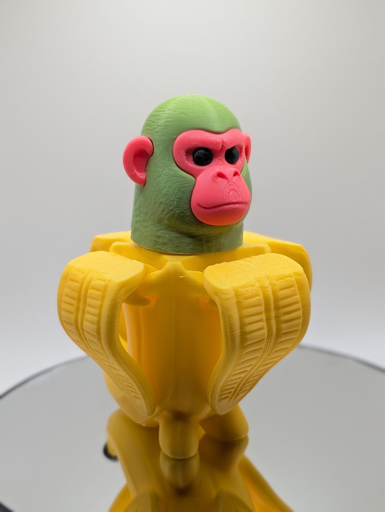 Puede incluir: Una escultura impresa en 3D de un mono con cabeza verde, cara y orejas rosas y ojos negros. El mono lleva un traje amarillo con forma de pl&aacute;tano. La escultura est&aacute; sobre una superficie reflectante.