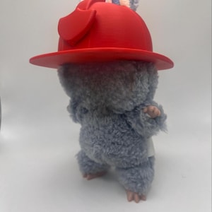 May include: A soft toy rodent wearing a vibrant red hat. The toy has grey fur, pink paws, and small ears. The hat is a hard, moulded plastic with a wide brim.