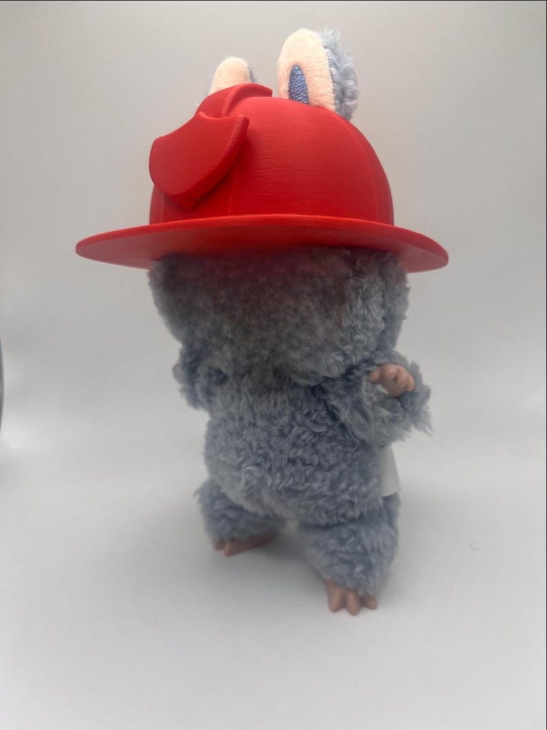 May include: A soft toy rodent wearing a vibrant red hat. The toy has grey fur, pink paws, and small ears. The hat is a hard, moulded plastic with a wide brim.