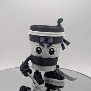 Puede incluir: Figura de taza de caf&eacute; con tem&aacute;tica ninja en blanco, negro y gris. La taza tiene una diadema negra con un emblema plateado, guantes negros y zapatillas deportivas. Sostiene dos espadas plateadas. La figura est&aacute; sobre una superficie reflectante.