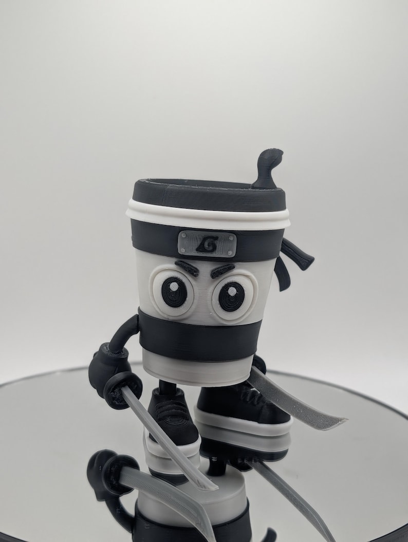 Puede incluir: Figura de taza de caf&eacute; con tem&aacute;tica ninja en blanco, negro y gris. La taza tiene una diadema negra con un emblema plateado, guantes negros y zapatillas deportivas. Sostiene dos espadas plateadas. La figura est&aacute; sobre una superficie reflectante.