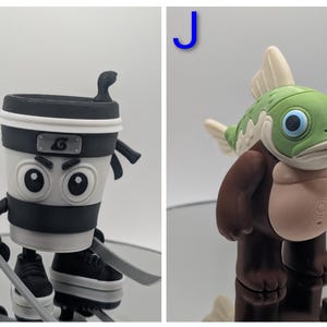 Puede incluir: Dos figuras caprichosas. Una es una taza de caf&eacute; ninja con una diadema negra, sosteniendo espadas y usando zapatillas. La otra es una criatura parecida a un pez con cabeza verde, aletas blancas y cuerpo marr&oacute;n.