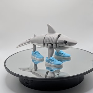 Puede incluir: Una figura de tibur&oacute;n articulada impresa en 3D en gris, con zapatillas deportivas en miniatura de color azul claro y blanco. El tibur&oacute;n est&aacute; posado sobre una superficie reflectante, mostrando su dise&ntilde;o y los peque&ntilde;os zapatos.