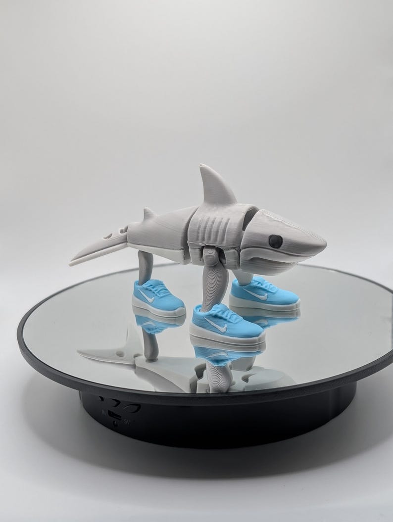 Puede incluir: Una figura de tibur&oacute;n articulada impresa en 3D en gris, con zapatillas deportivas en miniatura de color azul claro y blanco. El tibur&oacute;n est&aacute; posado sobre una superficie reflectante, mostrando su dise&ntilde;o y los peque&ntilde;os zapatos.