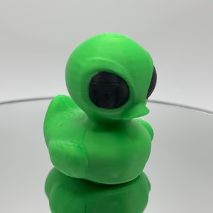 May include: A green rubber duck with black eyes, shaped like an alien. The duck is sitting on a reflective surface.