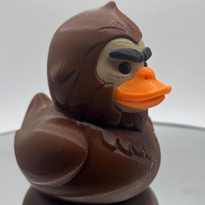 May include: A brown rubber duck with a Sasquatch-like head and an orange beak. The duck has a textured surface and is sitting on a reflective surface. The duck's eyes are black, and the face is tan.