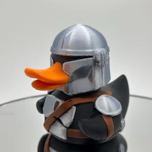 May include: A black rubber duck with a silver helmet and orange beak. The duck is wearing a brown and silver armor.