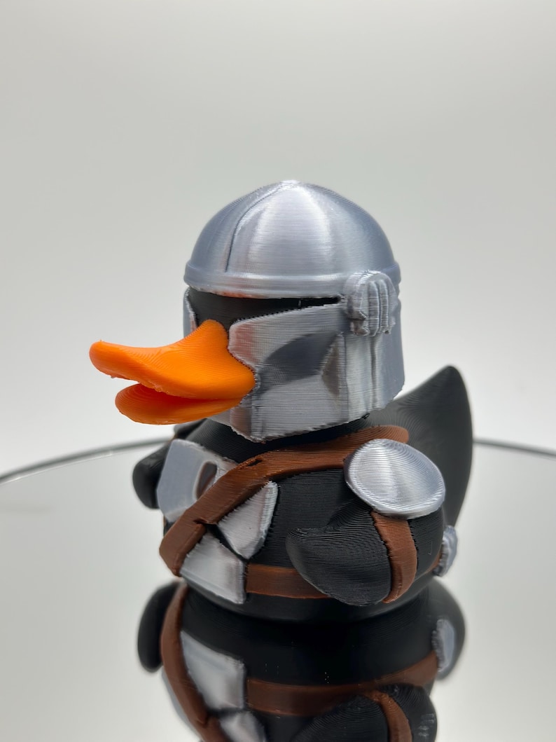 May include: A black rubber duck with a silver helmet and orange beak. The duck is wearing a brown and silver armor.