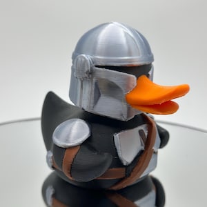 May include: A black rubber duck with a silver helmet and orange beak. The duck is wearing a brown belt and silver armor.