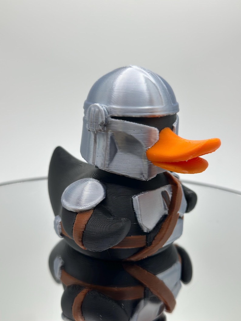 May include: A black rubber duck with a silver helmet and orange beak. The duck is wearing a brown belt and silver armor.