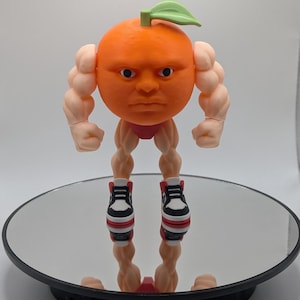 Puede incluir: Figura de naranja impresa en 3D con rostro humano, brazos y piernas musculosos. La naranja tiene una hoja verde y lleva calzoncillos rojos y zapatillas deportivas blancas y negras. La figura est&aacute; sobre una superficie reflectante.